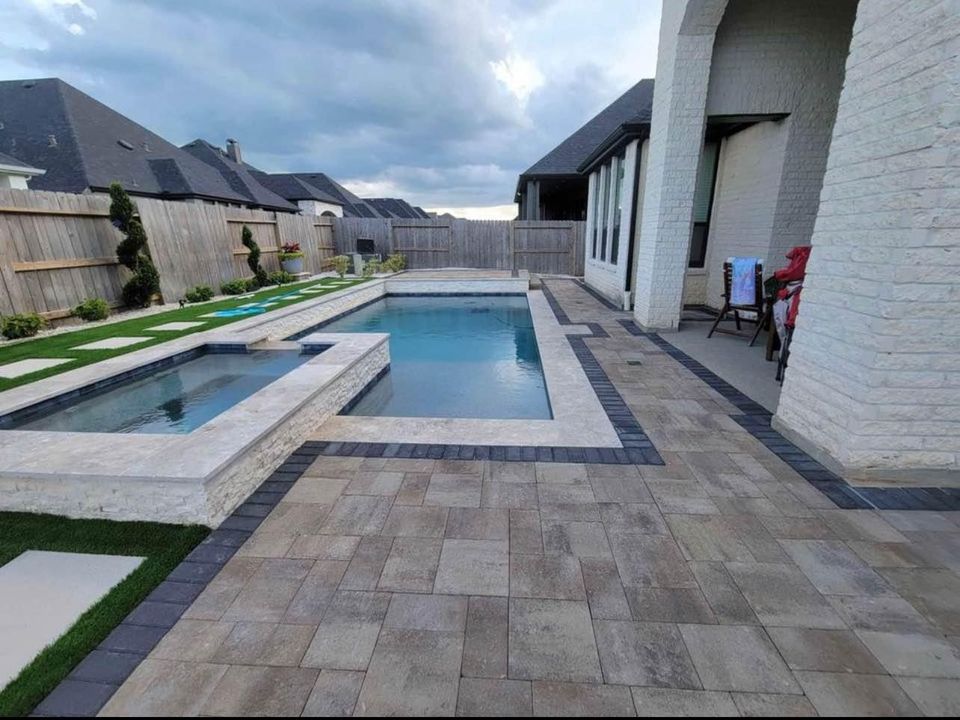 A modern backyard pool area features a rectangular swimming pool and adjacent hot tub, elegantly surrounded by expertly installed stone pavers. Nearby, a green lawn complements the stylish setting, all under a cloudy sky with several houses in the background.