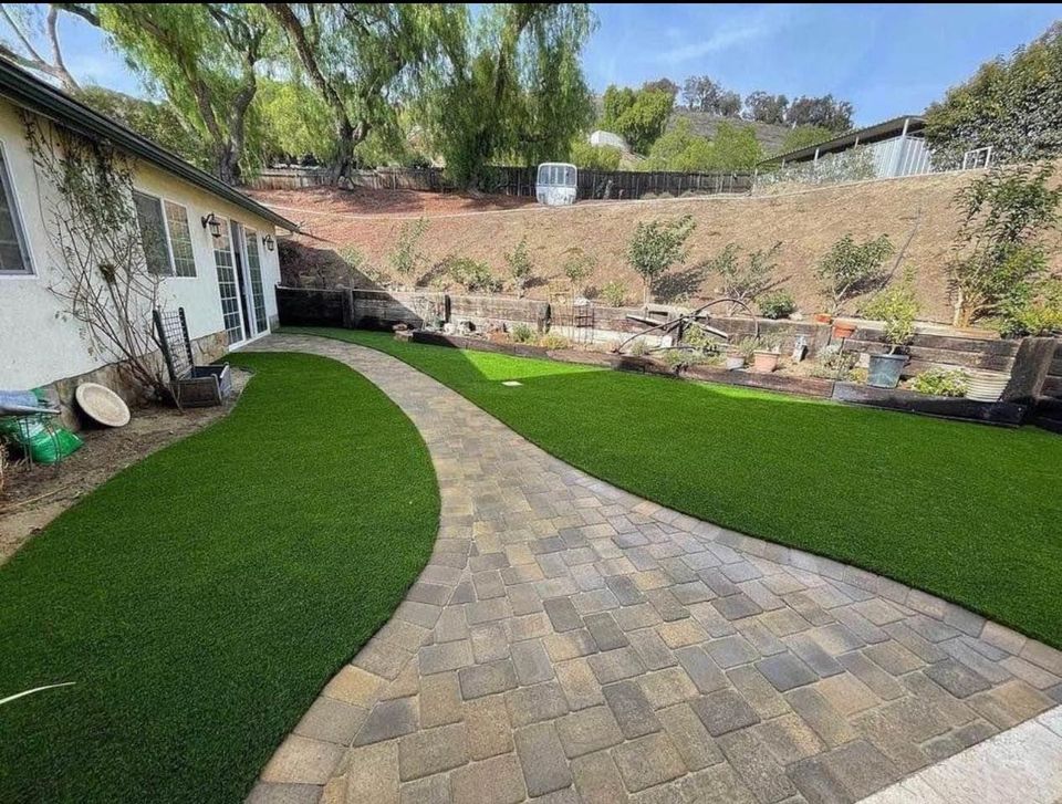 A backyard with a large, curved stone path, expertly crafted through paver installation, leading from a house to a garden area. The yard features neatly trimmed artificial grass on both sides of the path, with various plants and garden beds along a hillside.